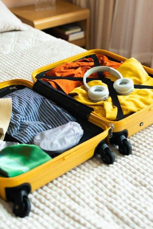 Open yellow suitcase with different family clothes and accessories on bed in light bedroom