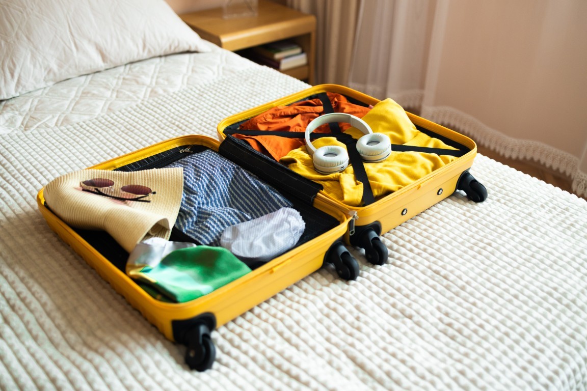 Open yellow suitcase with different family clothes and accessories on bed in light bedroom